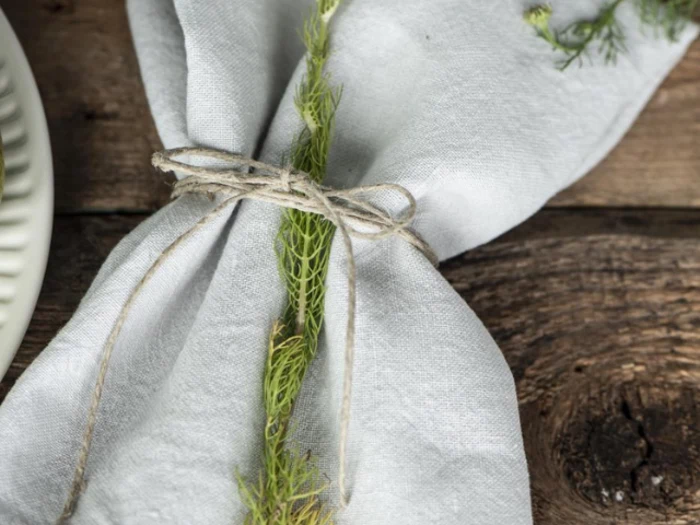 En hvid stofserviet bundet med sejlgarn og en kvist grøn urt, placeret på et rustikt træbord.