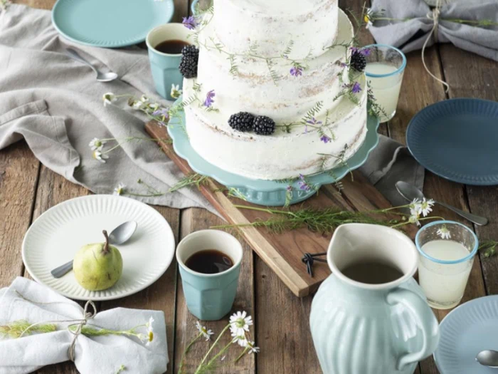 Et rustikt bord dækket med kaffe, mælk, en pære, blå tallerkener, servietter og en nøgen kage i to etager pyntet med brombær og blomster.