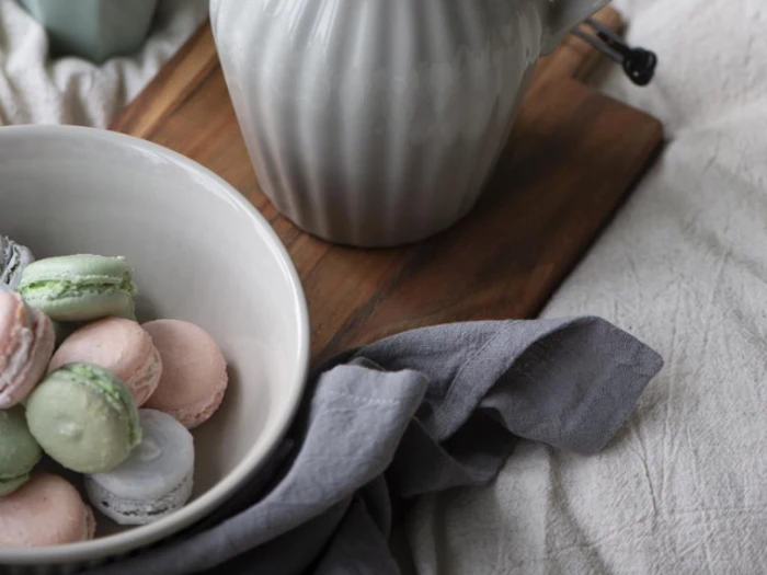 En hvid skål med pastelfarvede macarons står på en træplade ved siden af en grå tekande og en grå klud på en lys stofoverflade.