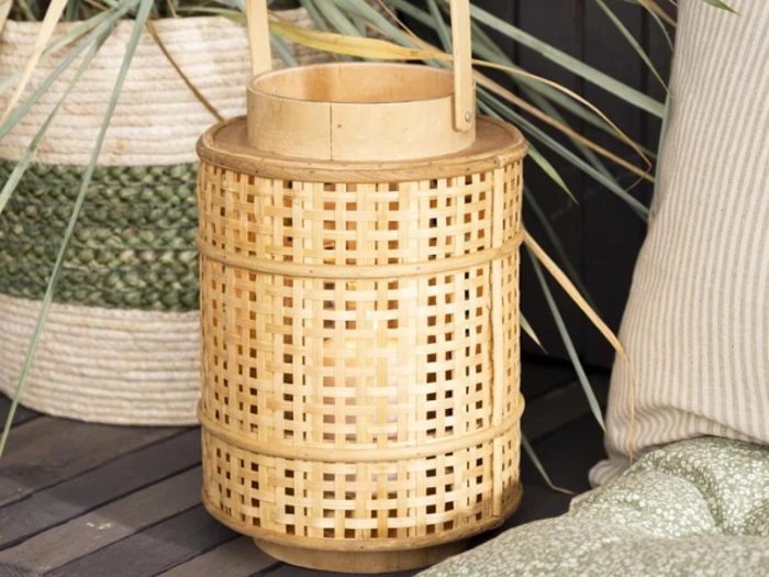 En flettet bambuslygte med hank står på en træplade ved siden af en grøn og hvid kurv og en stribet pude.