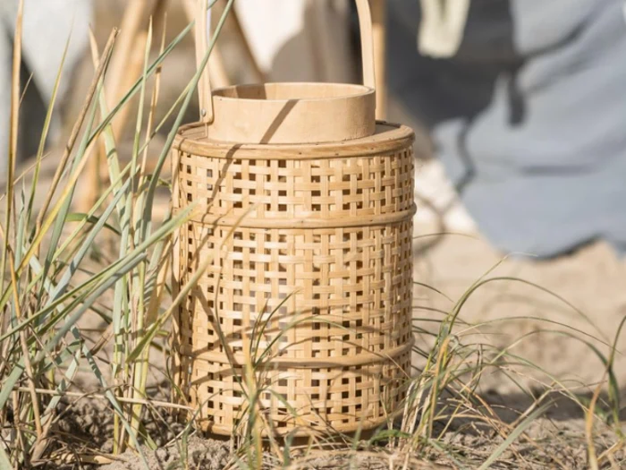 En flettet lanterne står på sandjord blandt højt græs med slørede former i baggrunden.