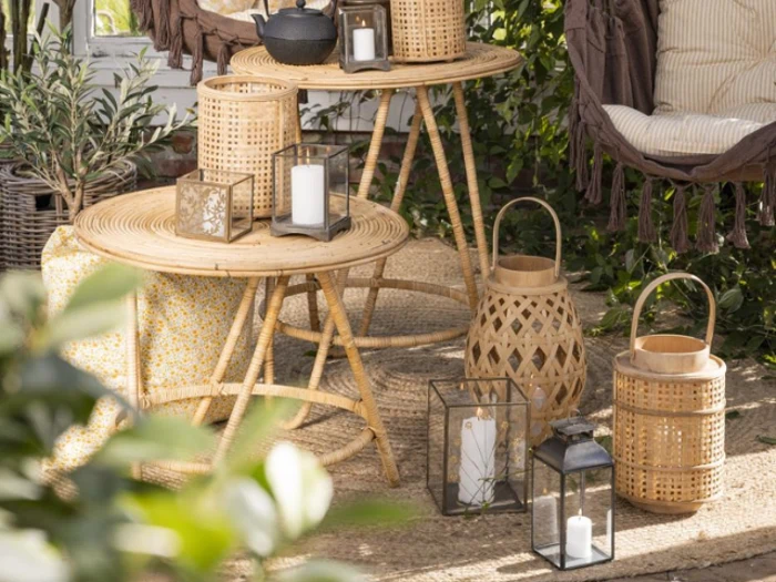 Rattanborde og lanterner med stearinlys arrangeret udendørs, omgivet af grønne planter, puder og en hyggelig hængestol, der skaber en varm og indbydende atmosfære.