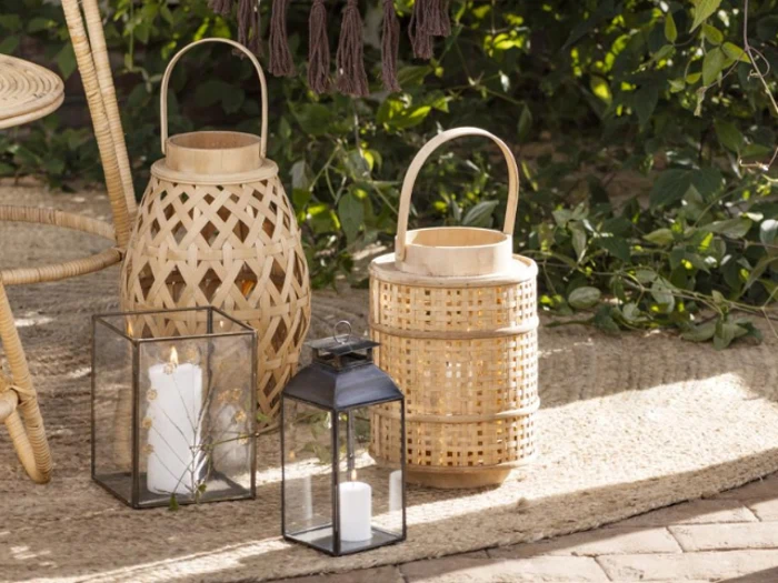 Tre lanterner - to af flettet bambus og en af sort metal - står på et tekstureret tæppe udendørs, hver med et stearinlys, omgivet af grønt og naturligt lys.