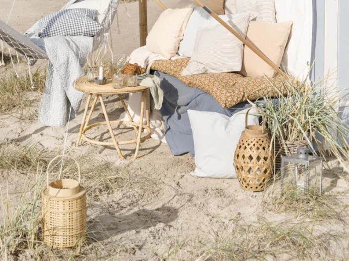 Hyggelig strandopsætning med hynder, tæpper, et lille kurvebord, lanterner og tørret græs på sandjord i blødt sollys.