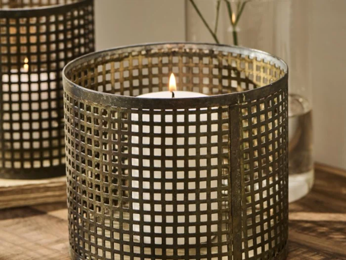 Et tændt hvidt stearinlys sidder i en metalnetholder på en træoverflade med en glasvase i baggrunden.