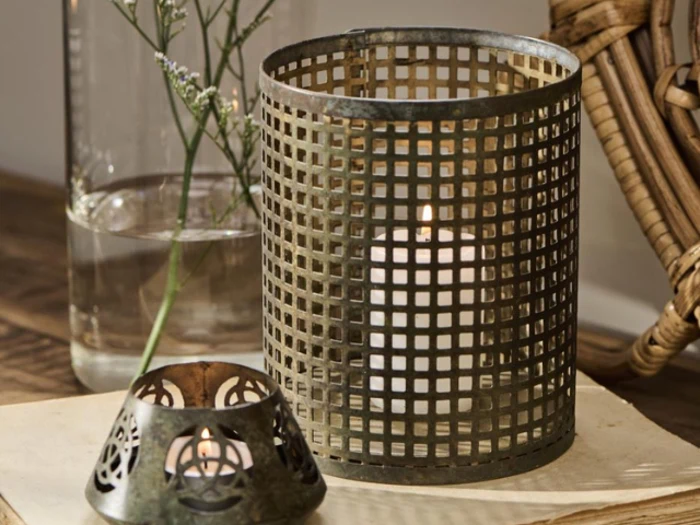 Et tændt hvidt stearinlys i en metalgitterholder står ved siden af en lille dekorativ fyrfadsstage og en glasvase med vand og grønt på en træplade.