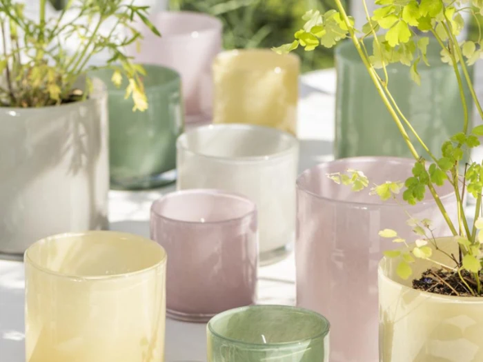 En gruppe pastelfarvede glaspotter, hvoraf nogle indeholder grønne planter, arrangeret på en solbeskinnet hvid overflade udendørs.