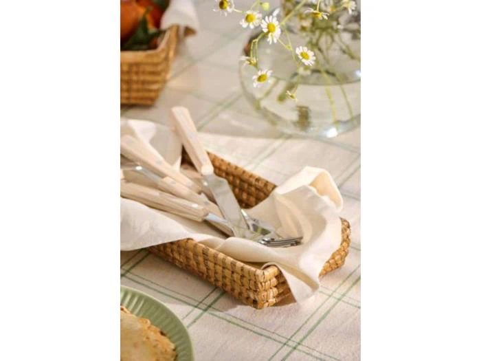 En flettet kurv foret med en hvid serviet rummer sølvtøj på en ternet dug, og der er blomster i en glasvase i nærheden.