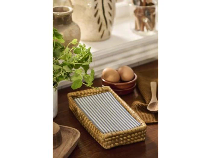 En flettet kurv med stribede servietter står på et træbord med en plante, keramikkrukker, en skål med æg og træredskaber i nærheden.