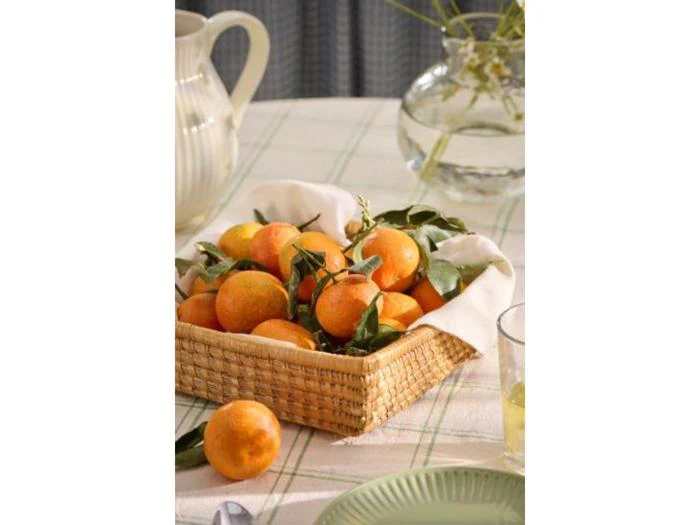 En flettet kurv fyldt med friske appelsiner og grønne blade står på et bord med en ternet dug ved siden af en glaskande og en vase med vand og stilke.