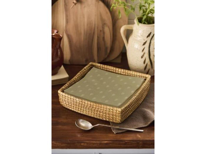 En firkantet kurv med grønne servietter står på et træbord ved siden af en ske, en tekstureret kande og skærebrætter af træ.