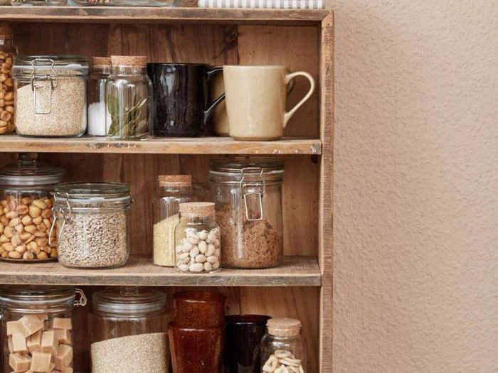 Træhylder med glaskrukker med tørvarer, et beige krus og andre ting til spisekammeret mod en struktureret beige væg.