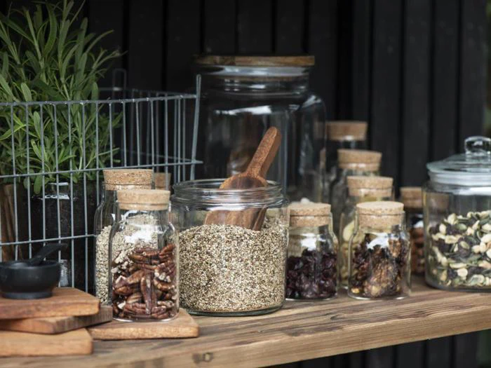 Flere glaskrukker med korklåg fyldt med tørrede fødevarer og frø står på en træhylde med en potteplante og en trådkurv i baggrunden.