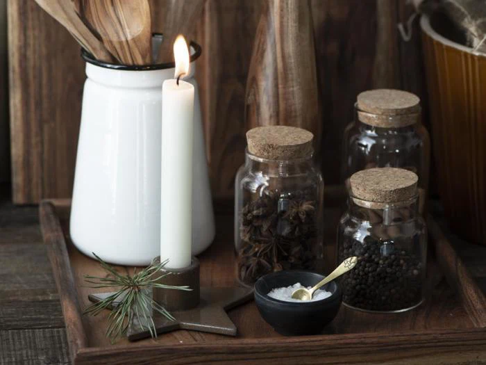 Et tændt hvidt lys, tre krydderikrukker, en hvid kop med træredskaber og et lille fad med en ske står på en træbakke. Fyrrekviste pynter på bakken.
