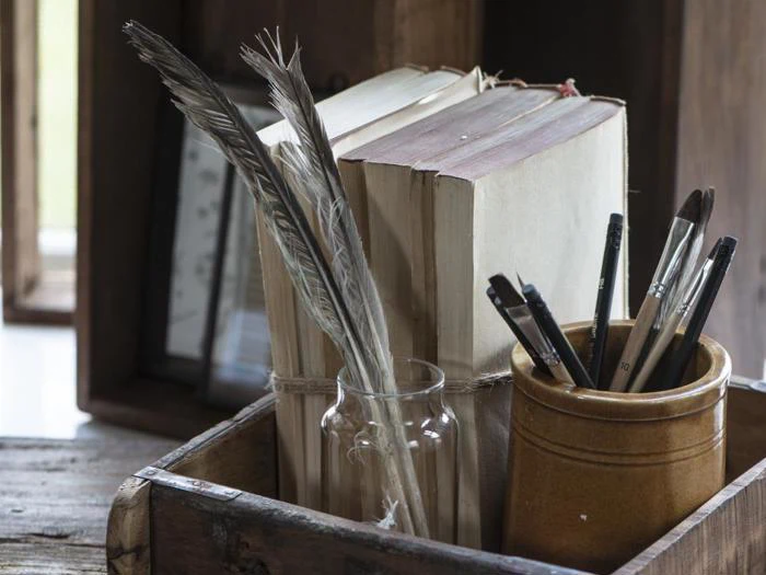 En trækasse indeholder gamle bøger, en glaskrukke med fjer og en keramikkop med pensler på en rustik træoverflade.