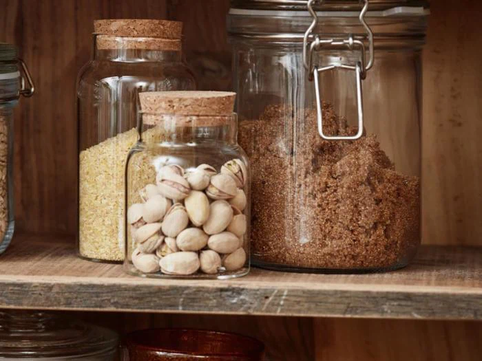 Tre glaskrukker med korklåg på en træhylde med brunt sukker, hakkede nødder og afskallede pistacienødder.