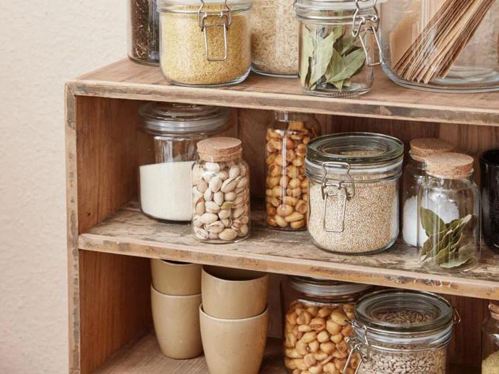Glaskrukker fyldt med korn, nødder og krydderier er pænt arrangeret på træhylder med stablede beige kopper på den nederste hylde. Opstillingen ser ryddelig og organiseret ud.