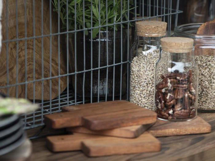 Tre små skærebrætter af træ er stablet ved siden af glaskrukker fyldt med kerner og nødder foran en trådhylde og en potteplante.