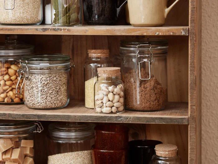 Glaskrukker fyldt med forskellige spisekammervarer som nødder, frø, pistacienødder og brun farin er sirligt anbragt på træhylder.