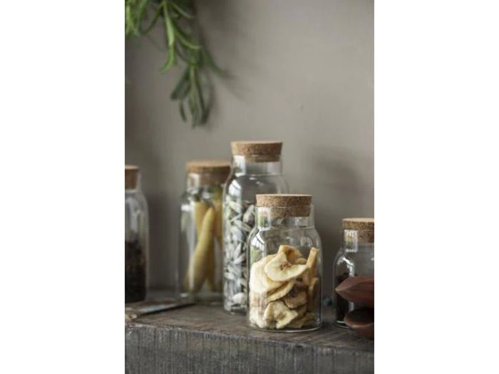 Glaskrukker med korklåg fyldt med tørrede bananchips, frø og andre konserverede fødevarer står på en rustik træhylde med nogle grønne krydderurter hængende i nærheden.