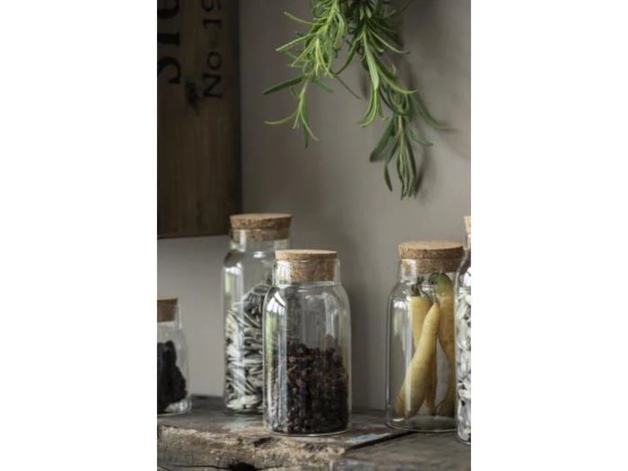 På en rustik træhylde står glaskrukker med korklåg, som indeholder forskellige tørrede frø, krydderier og bønner, og over dem hænger et bundt friske krydderurter.