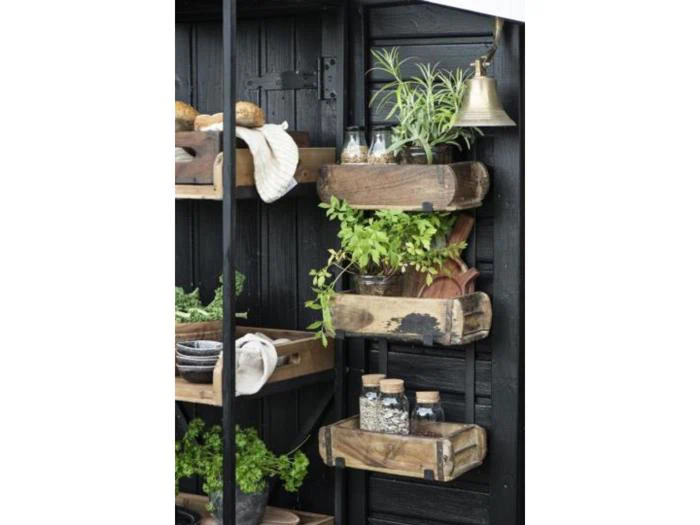 Træhylder på en sort væg rummer krydderurter, krukker og køkkenting ved siden af en messingklokke i et rustikt køkken- eller havemiljø.