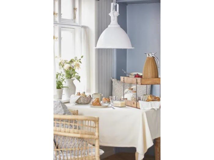 En hyggelig spiseplads med hvid dug, morgenmadsprodukter, kurvestol, blomster i en vase og en stor hvid pendellampe over bordet nær et vindue.