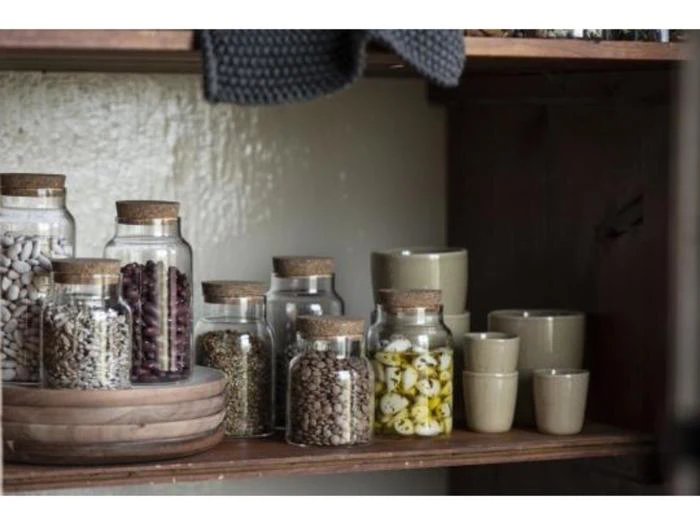 Glaskrukker med korklåg med forskellige bønner og krydderier står på en træhylde sammen med stablede keramiske kopper og skåle.