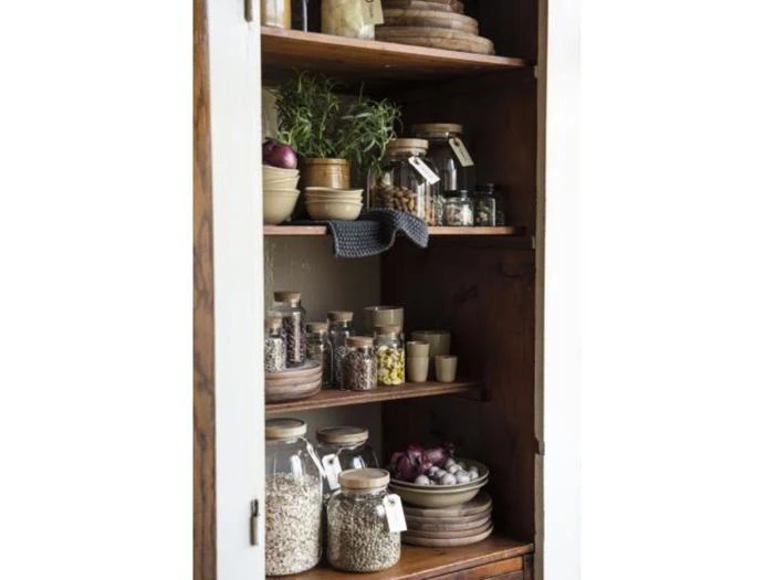 Et spisekammerskab i træ med hylder med krukker med korn, krydderier, tørrede fødevarer, krydderurter og skåle, alt sammen sirligt organiseret.