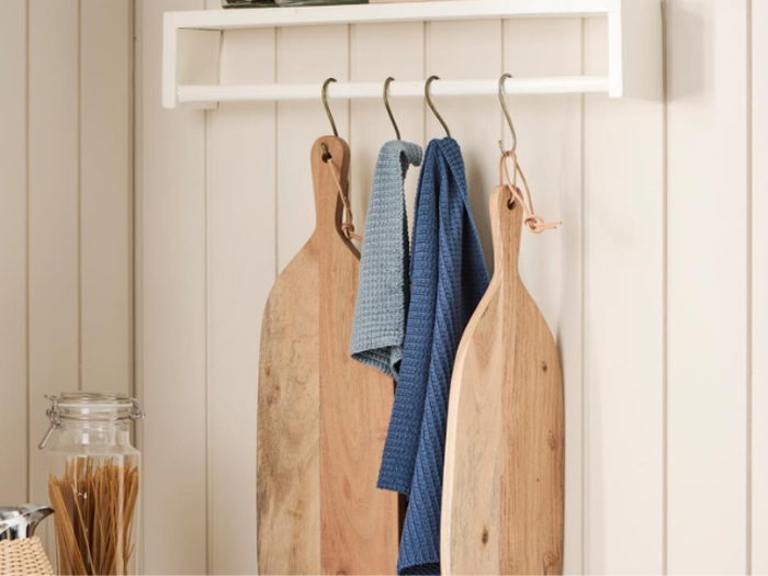 To skærebrætter af træ og to blå viskestykker hænger på kroge på et hvidt stativ, der er monteret på en væg med lyse paneler. Et glas spaghetti står på køkkenbordet nedenunder.