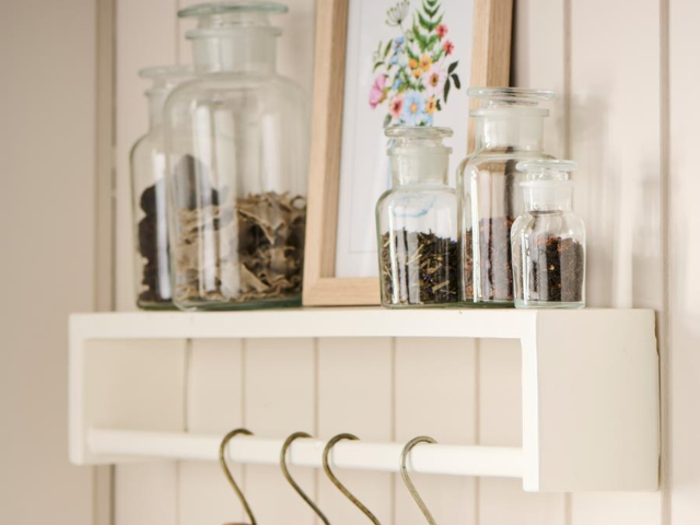 En hvid væghylde rummer glaskrukker med tørrede krydderurter eller krydderier, et indrammet blomsterprint og har flere metalkroge hængende under sig.