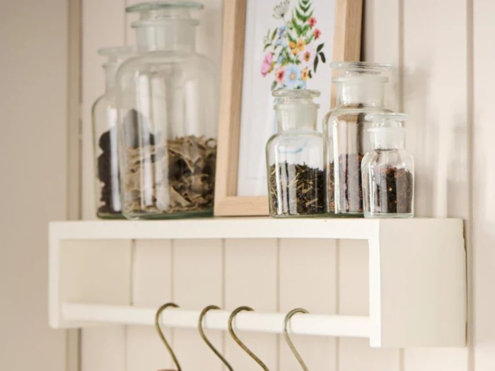 En hvid væghylde rummer glaskrukker med tørrede krydderurter eller krydderier, et indrammet blomsterprint og har flere metalkroge hængende under sig.