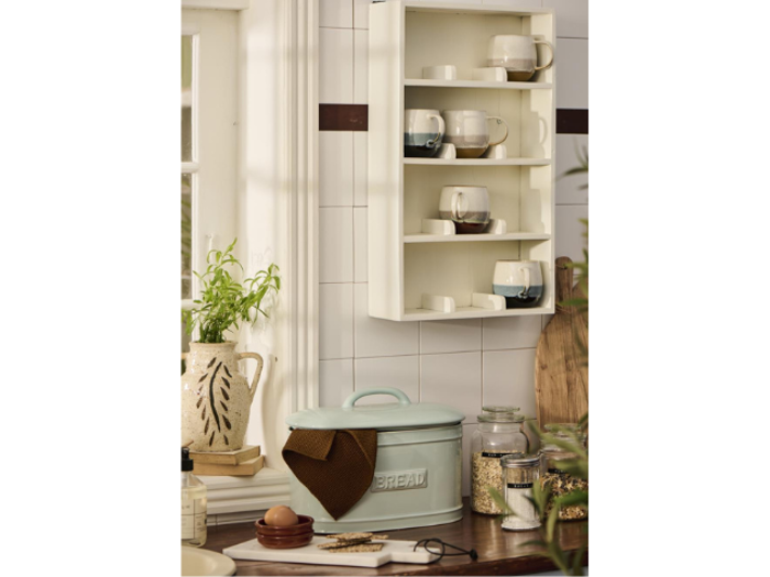 En hyggelig køkkenscene med en hvid hylde med forskellige krus, en lyseblå brødkasse med et brunt klæde, krukker, en plante og sollys, der strømmer ind gennem et vindue.