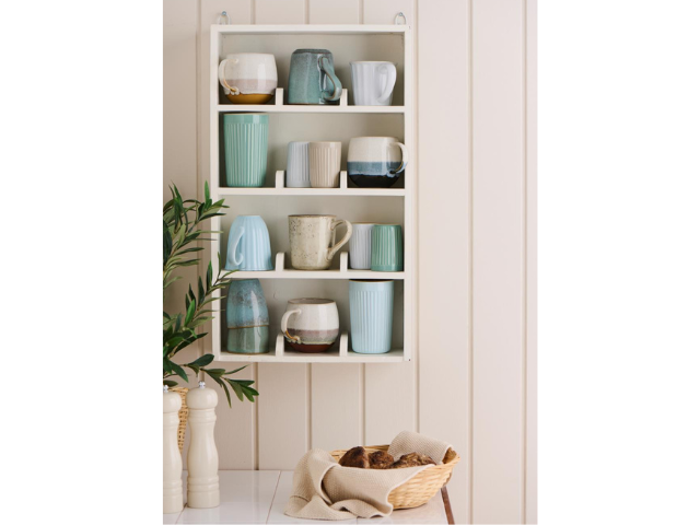 En hvid væghængt hylde viser forskellige keramikkrus i bløde farver. Nedenfor står en kurv med brød ved siden af en plante og to hvide peber- og saltkværne på et bord.