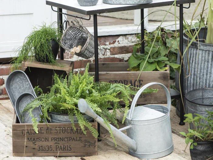 Haveredskaber af metal, vandkande, potteplanter og opbevaringskasser af træ er arrangeret udendørs nær en murstensvæg og en hvid struktur.