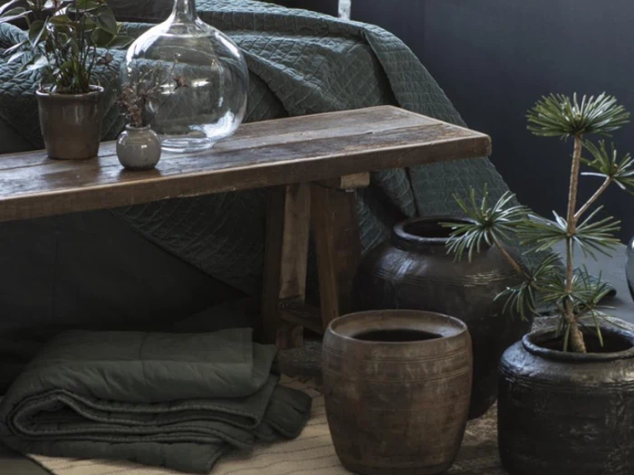 En rustik træbænk med potteplanter, glaskrukke og keramikpotter står ved siden af et foldet tæppe og quiltet sengetøj i jordfarver.