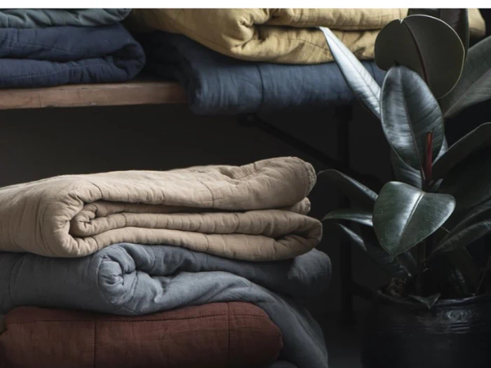 Stabler af foldede tæpper i beige, blå og rødbrune nuancer står ved siden af en potteplante med blanke, mørkegrønne blade.