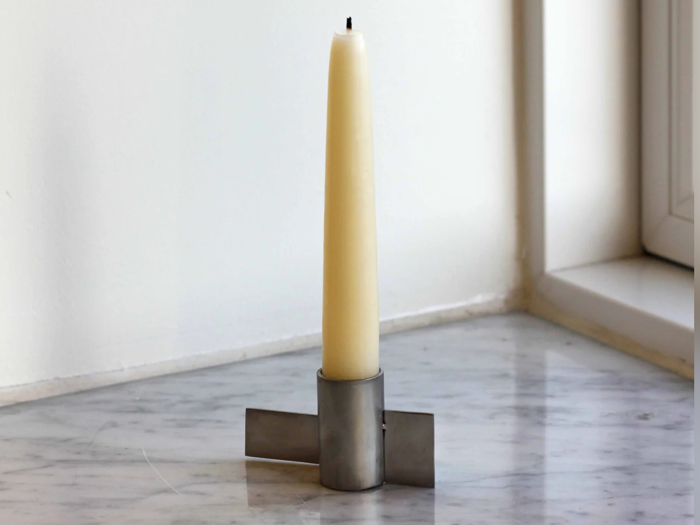 Et højt, cremefarvet stearinlys står oprejst i en moderne, geometrisk sølvholder på en marmorflade nær et vindue.