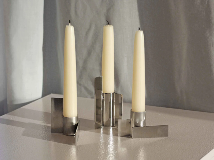 Tre utændte cremefarvede kertelys står i moderne, geometriske sølvholdere på en hvid struktureret overflade med et blødt gråt gardin som baggrund.