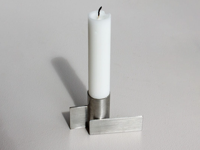Et hvidt stearinlys står oprejst i en moderne, sølvfarvet metalholder med geometriske, rektangulære former og kaster en svag skygge på en lys overflade.