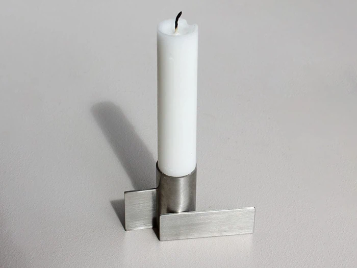Et hvidt stearinlys står oprejst i en moderne, sølvfarvet metalholder med geometriske, rektangulære former og kaster en svag skygge på en lys overflade.