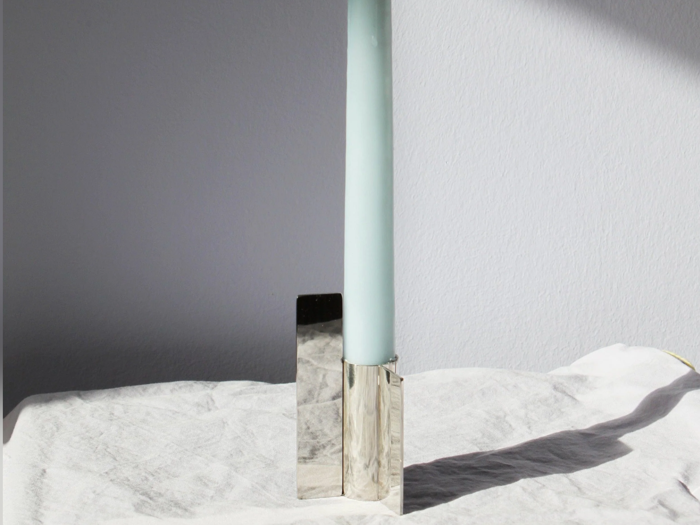 Et lyseblåt stearinlys står i en moderne, geometrisk sølvstage på et hvidt klæde, mens sollyset kaster skygger på væggen og stoffet.