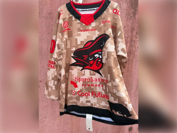 En beige hockeyt-shirt i camouflagemønster med et piratlogo, røde sponsorlogoer og teksten »Sparekassen Danmark« og »Cool Future«, der hænger foran en lyserød væg med struktur.