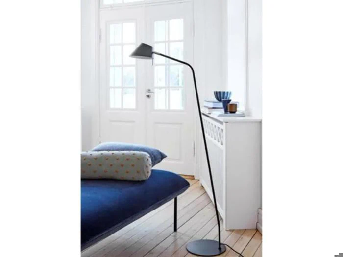 En moderne sort gulvlampe står ved siden af en blå sofa med mønstrede puder i et lyst rum med trægulv og store vinduer.