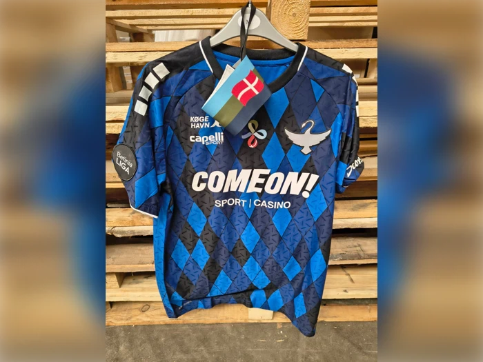 En blå og sort mønstret fodboldtrøje med sponsorlogoer hænger på en bøjle med et lille dansk flag fastgjort til den.