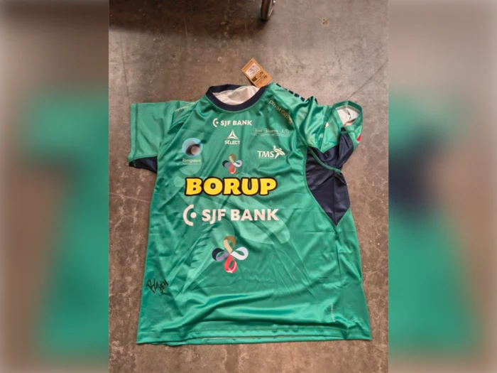 Grøn sportstrøje med korte ærmer, holdlogoer, sponsorer og en signatur nederst til venstre.