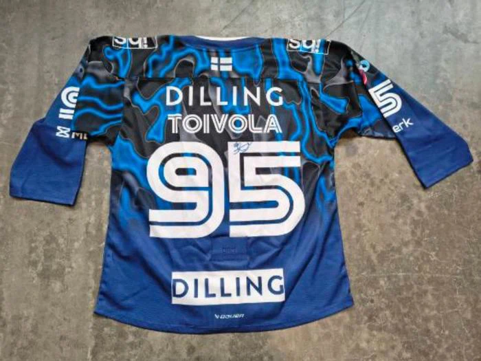 En blå og sort ishockeyskjorte med påskriften »DILLING«, »TOIVOLA« og nummeret 95 på ryggen, der ligger fladt på et betongulv.