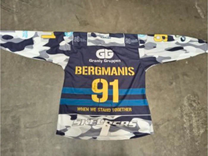En blå, hvid og grå hockeytrøje i camouflagemønster med teksten Bergmanis 91 og WHEN WE STAND TOGETHER trykt på ryggen, der ligger fladt på et betongulv.