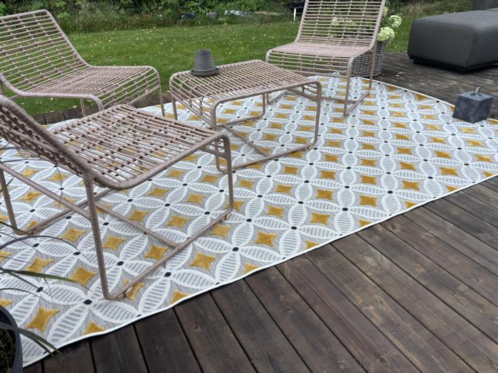 Tre kurvestole og et lille bord står på et mønstret udendørstæppe på en træterrasse, hvor man kan se grønt græs og planter i baggrunden.