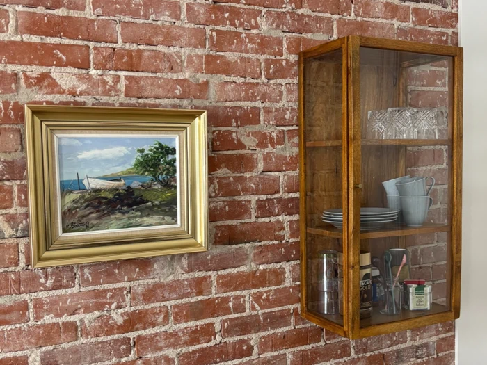 Et indrammet havmaleri hænger på en murstensvæg ved siden af et glasskab i træ med tallerkener, krus, glas og små krukker.